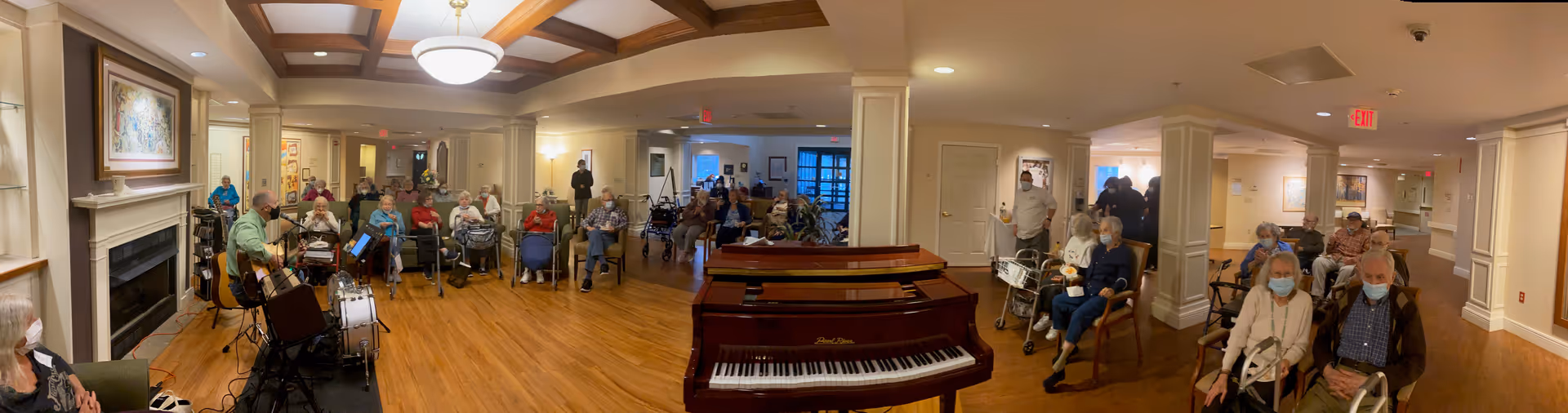 A group of elderly residents seated in a spacious common area of an assisted living facility, some wearing masks. A musician is playing guitar and drums on the left side near a fireplace, and a piano is centered in the room. The room has wooden floors, warm lighting, and a coffered ceiling.