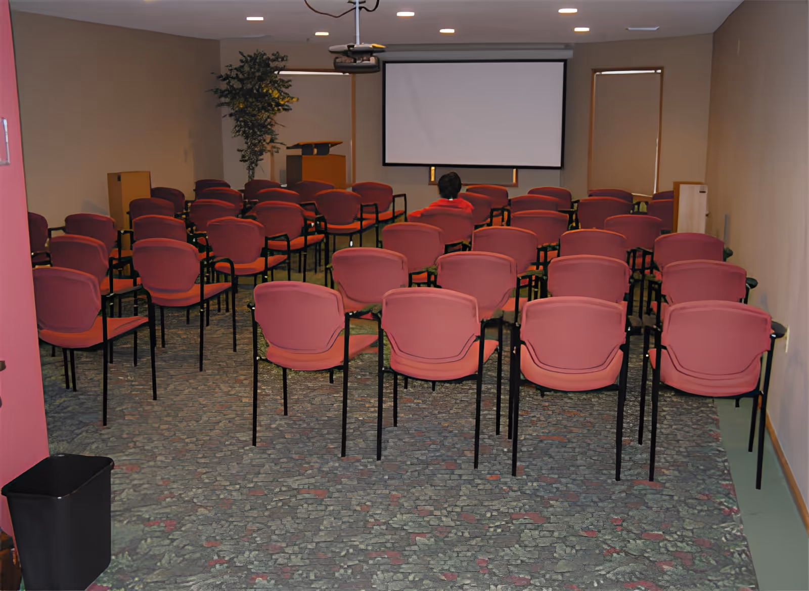 Rows of red chairs in a small meeting room facing a blank projection screen with one person seated near the front.
