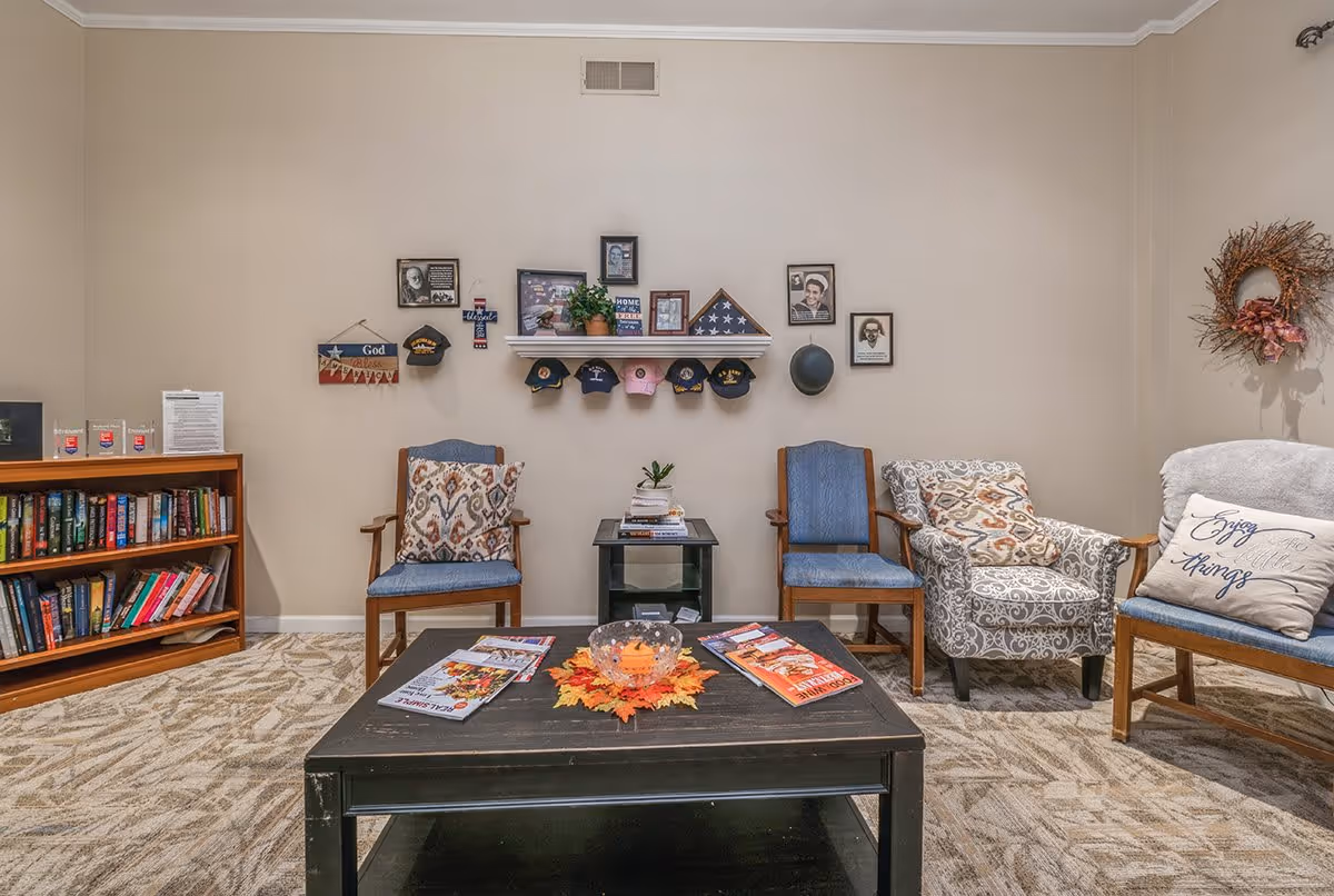Sitting area with upholstered and wooden chairs arranged around a coffee table, a bookshelf to the left, and a wall display of photos, hats, and decor.