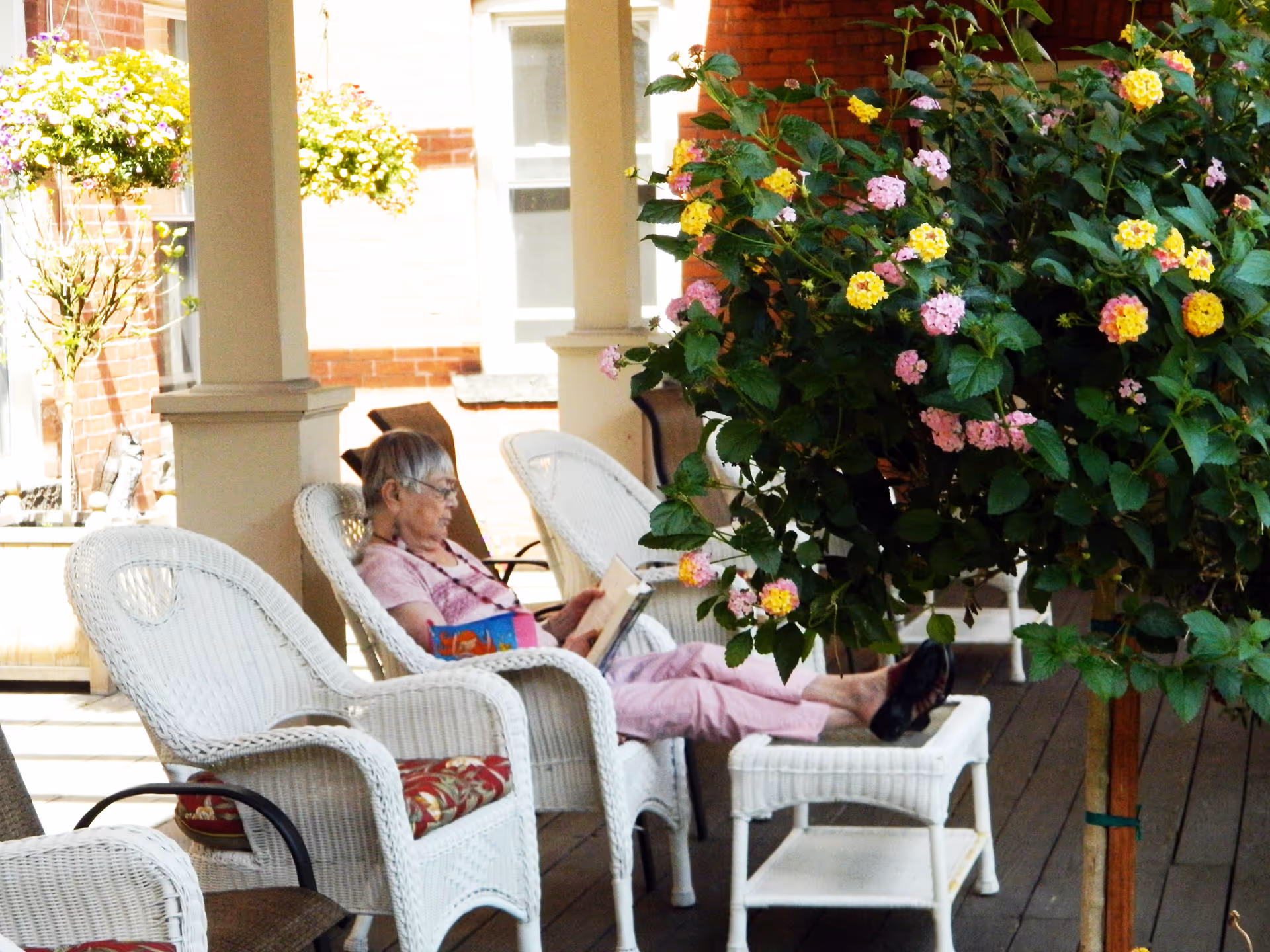 An elderly woman sitting on a white wicker chair on a covered porch, reading a book with her feet resting on a matching wicker footstool. The porch has wooden flooring and is decorated with hanging flower baskets and a large flowering plant with pink and yellow blooms nearby.