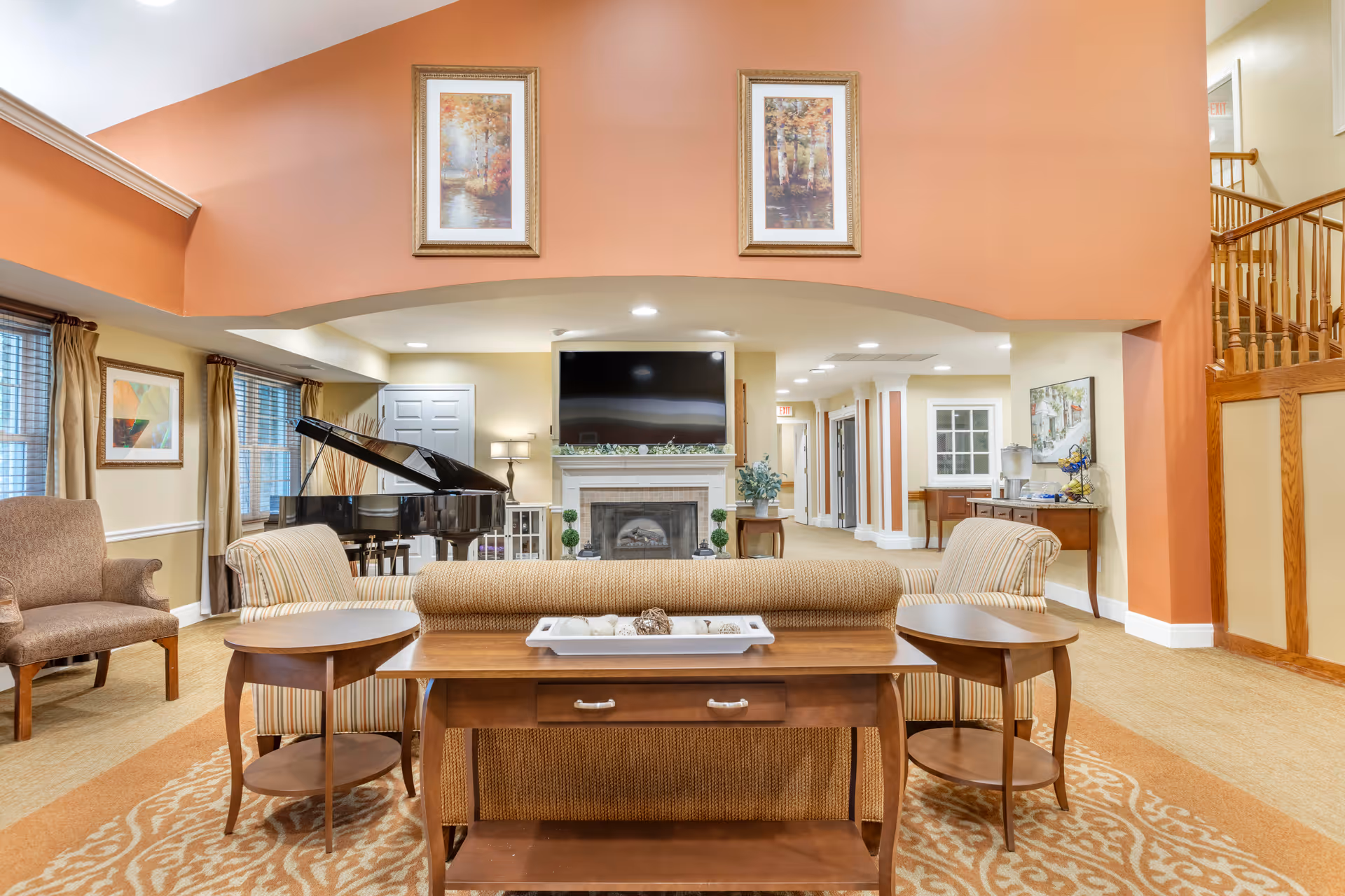 A spacious and well-lit living room area in an assisted living facility featuring a grand piano, a fireplace with a large flat-screen TV mounted above it, comfortable seating including armchairs and a sofa, wooden side tables, and framed artwork on the walls. The room has warm tones with beige and orange walls, carpeted floors, and a staircase with wooden railings on the right side.