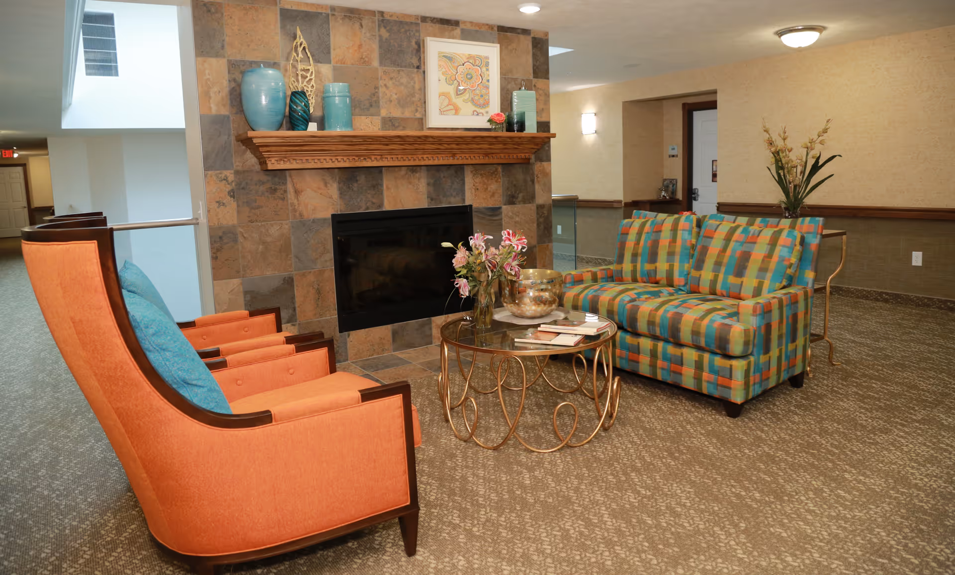A cozy seating area in a retirement resort featuring two orange armchairs with blue cushions, a colorful plaid loveseat, and a round glass coffee table with decorative items. The background includes a tiled fireplace with a wooden mantle adorned with vases and a framed picture, and a beige wall with a flower arrangement on a side table.