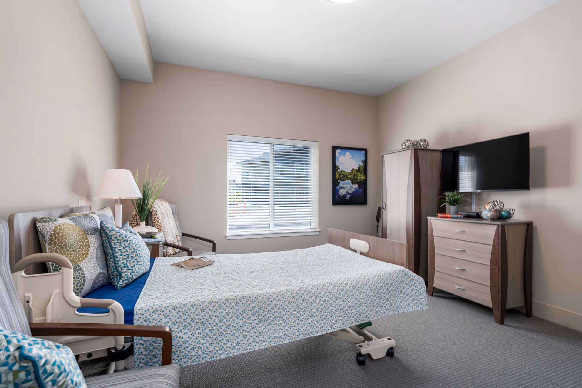 A tidy, well-lit rehabilitation bedroom with an adjustable hospital bed, patterned bedding, armchairs, a dresser with TV, and a window.