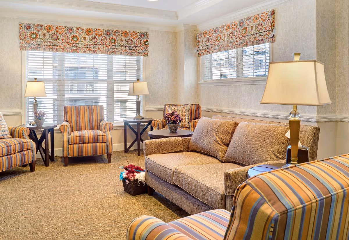 A cozy living room area with a beige sofa and several striped armchairs arranged around small wooden tables. The room has large windows with white blinds and floral valances, two table lamps providing warm light, and a basket with yarn and knitting needles on the floor.