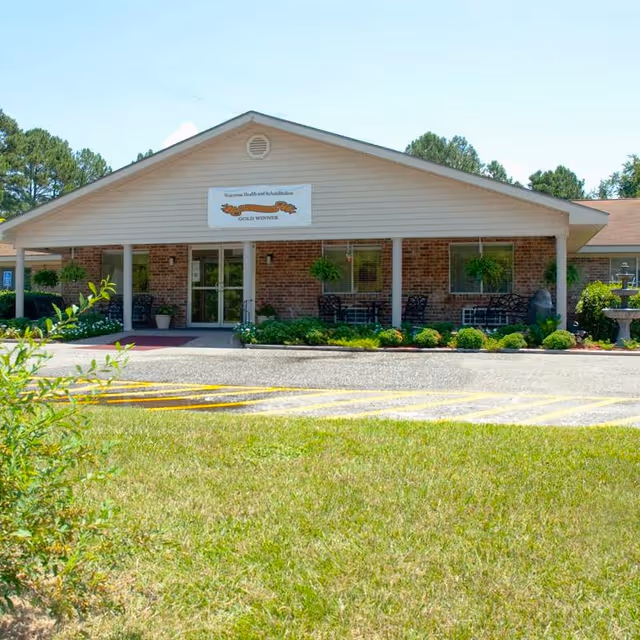 Front exterior view of Waycross Health and Rehabilitation building with a brick facade, a covered entrance, outdoor seating area with chairs and tables, hanging plants, and a well-maintained lawn and garden in front.