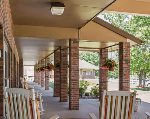 Covered outdoor patio area with brick columns and hanging flower pots. Several striped rocking chairs are placed along the walkway. Trees and a building are visible in the background under a clear sky.