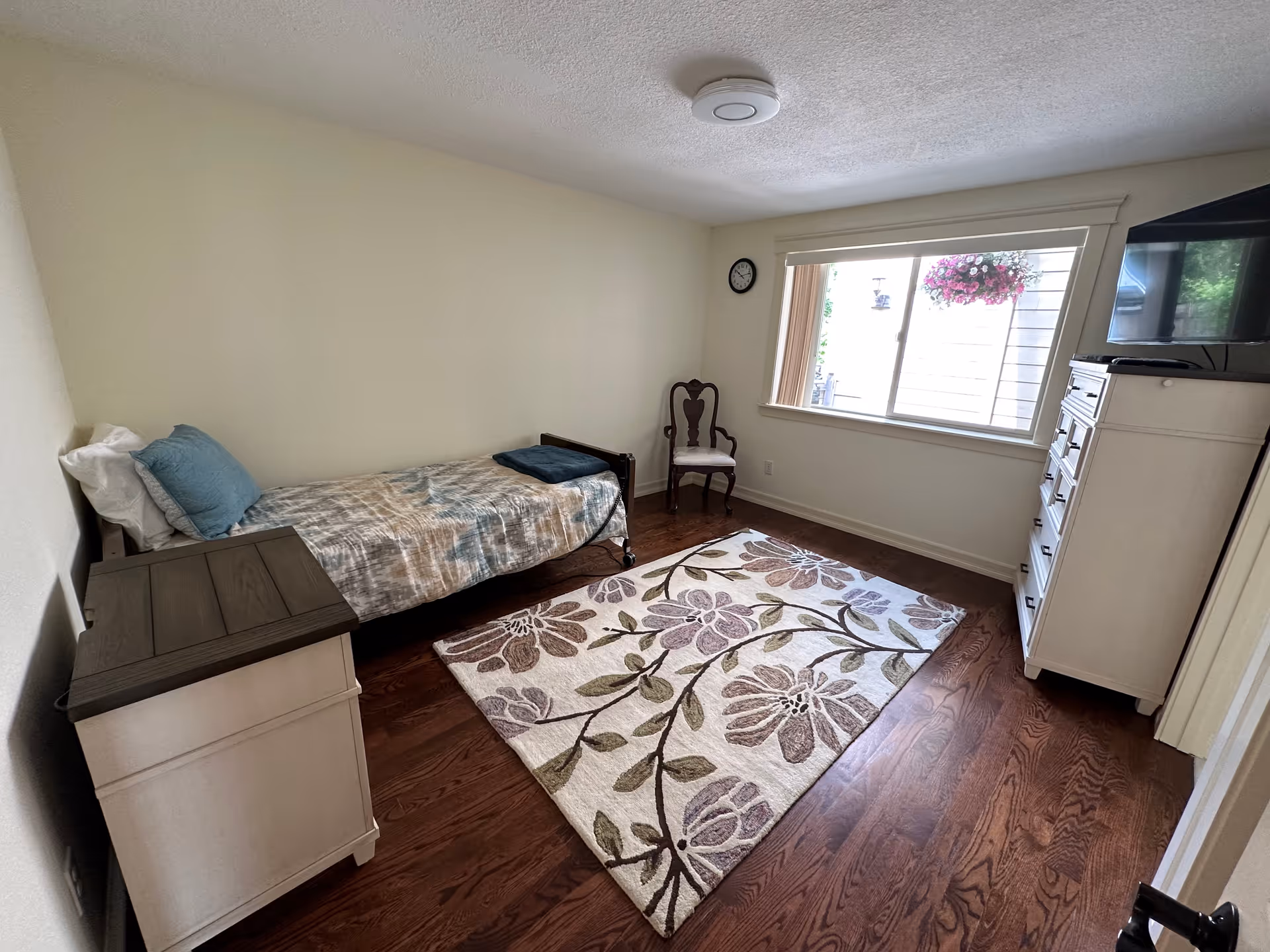 A small bedroom with a single bed against the left wall, a wooden nightstand next to the bed, a floral patterned area rug on the wooden floor, a wooden chair near a window with hanging flowers outside, a wall clock, and a white dresser with a TV on top.