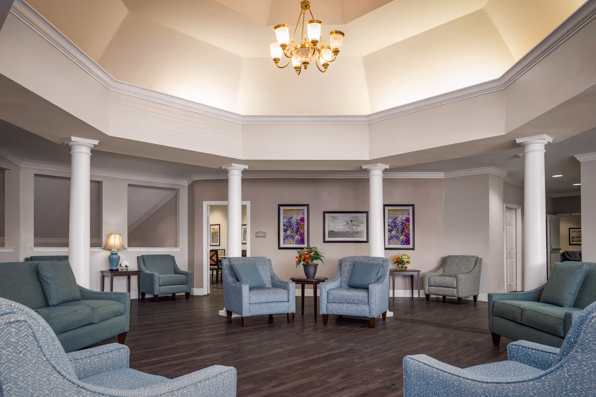 A spacious senior living facility common area with multiple blue upholstered armchairs arranged around small wooden tables with flower arrangements. The room features white columns, a high ceiling with a chandelier, and framed artwork on the walls. The flooring is dark wood, and there are doorways leading to other rooms.