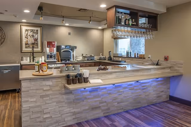 Modern kitchen area with a stone-textured counter and blue under-counter lighting. The counter has condiments, utensils, and napkins. Behind the counter are kitchen appliances including a coffee machine, toaster, and various bottles. There is a window with a view outside and a hanging rack with wine glasses above the counter.