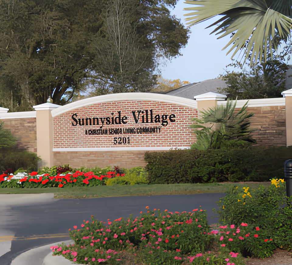 Brick entrance sign reading 'Sunnyside Village' set in landscaped grounds with flowers and trees.