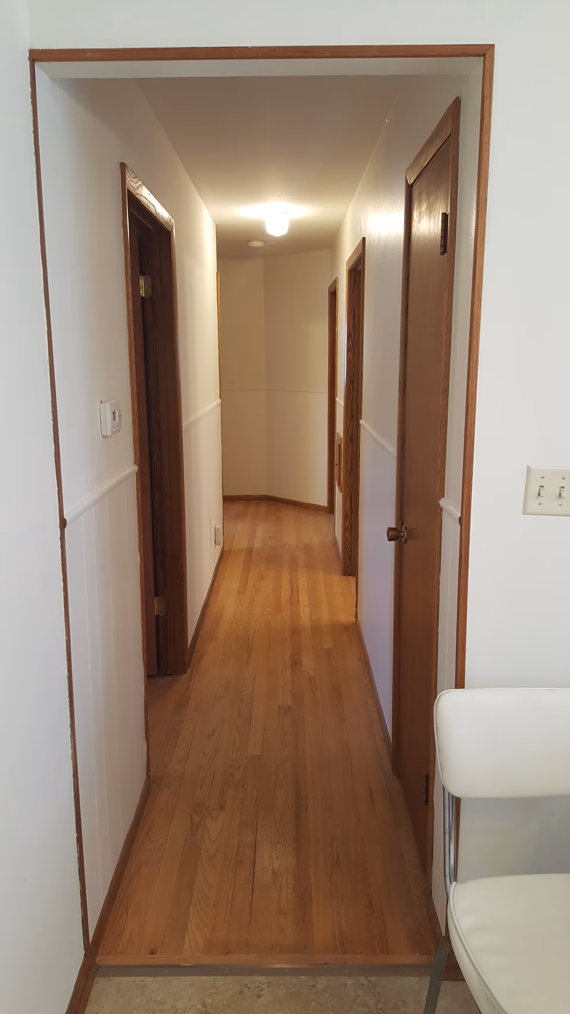 A narrow hallway with wooden floors and white walls featuring wooden door frames and doors on both sides. A ceiling light illuminates the hallway, and a white chair is partially visible on the right side near the entrance.