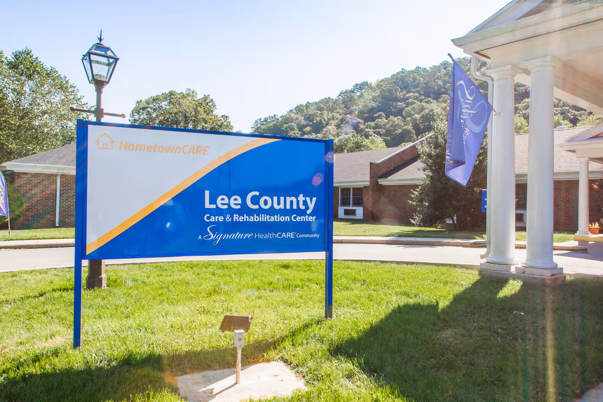 Outdoor view of the entrance area of Lee County Care & Rehabilitation Center with a large blue and white sign displaying the facility's name. The building is a single-story brick structure with white columns and a flagpole with a blue flag. Trees and hills are visible in the background under a clear sky.