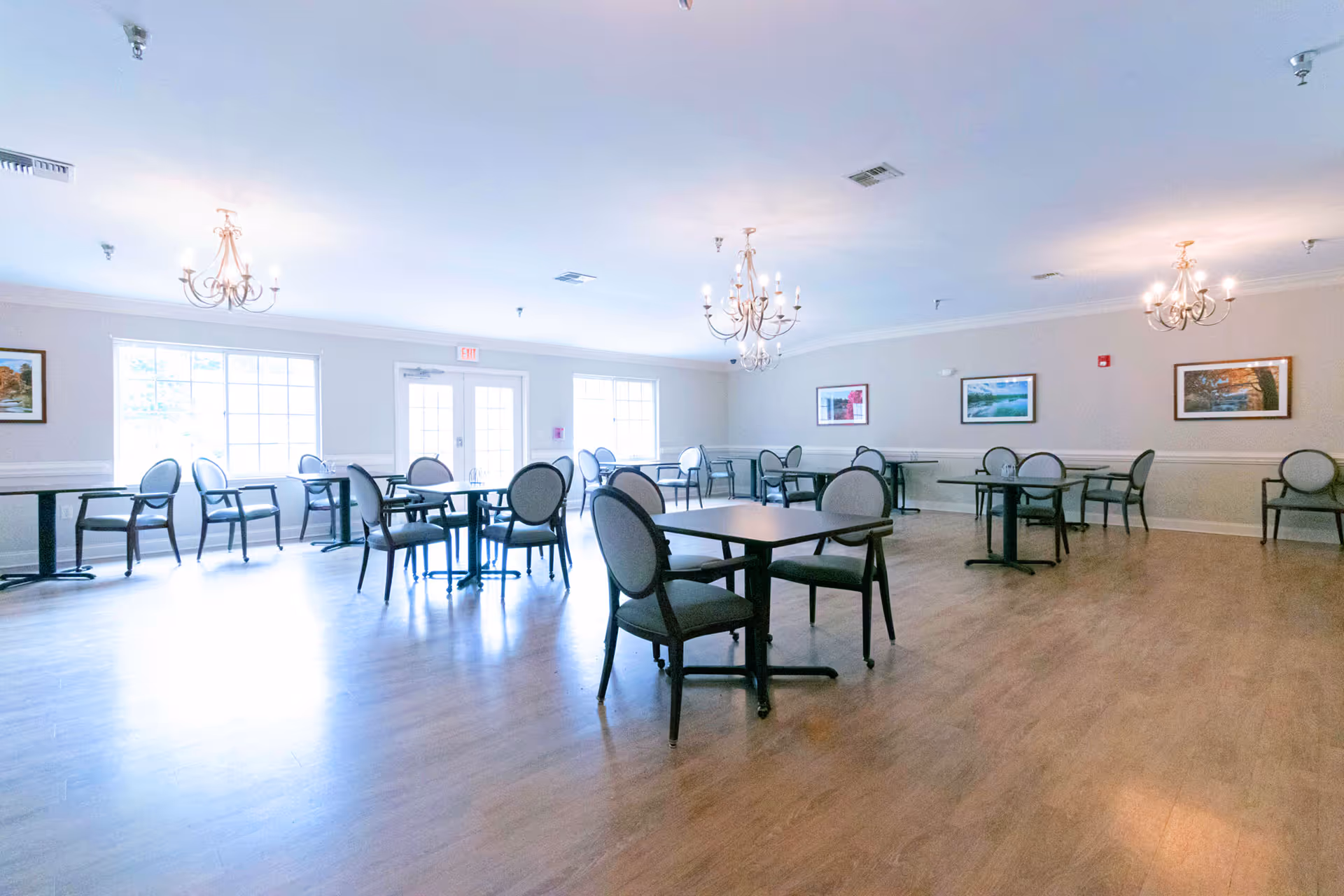 A large, well-lit senior living facility dining room with multiple tables and chairs, chandeliers, and wall art.
