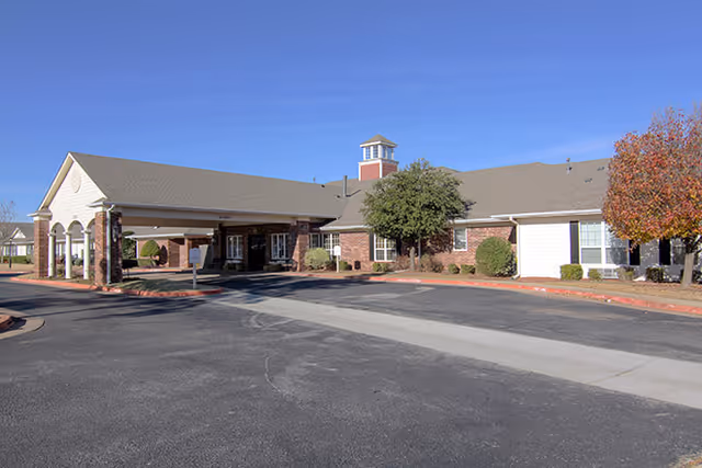 Exterior view of a single-story senior living facility building with a covered entrance, brick and siding walls, a small cupola on the roof, and landscaping including trees and shrubs under a clear blue sky.