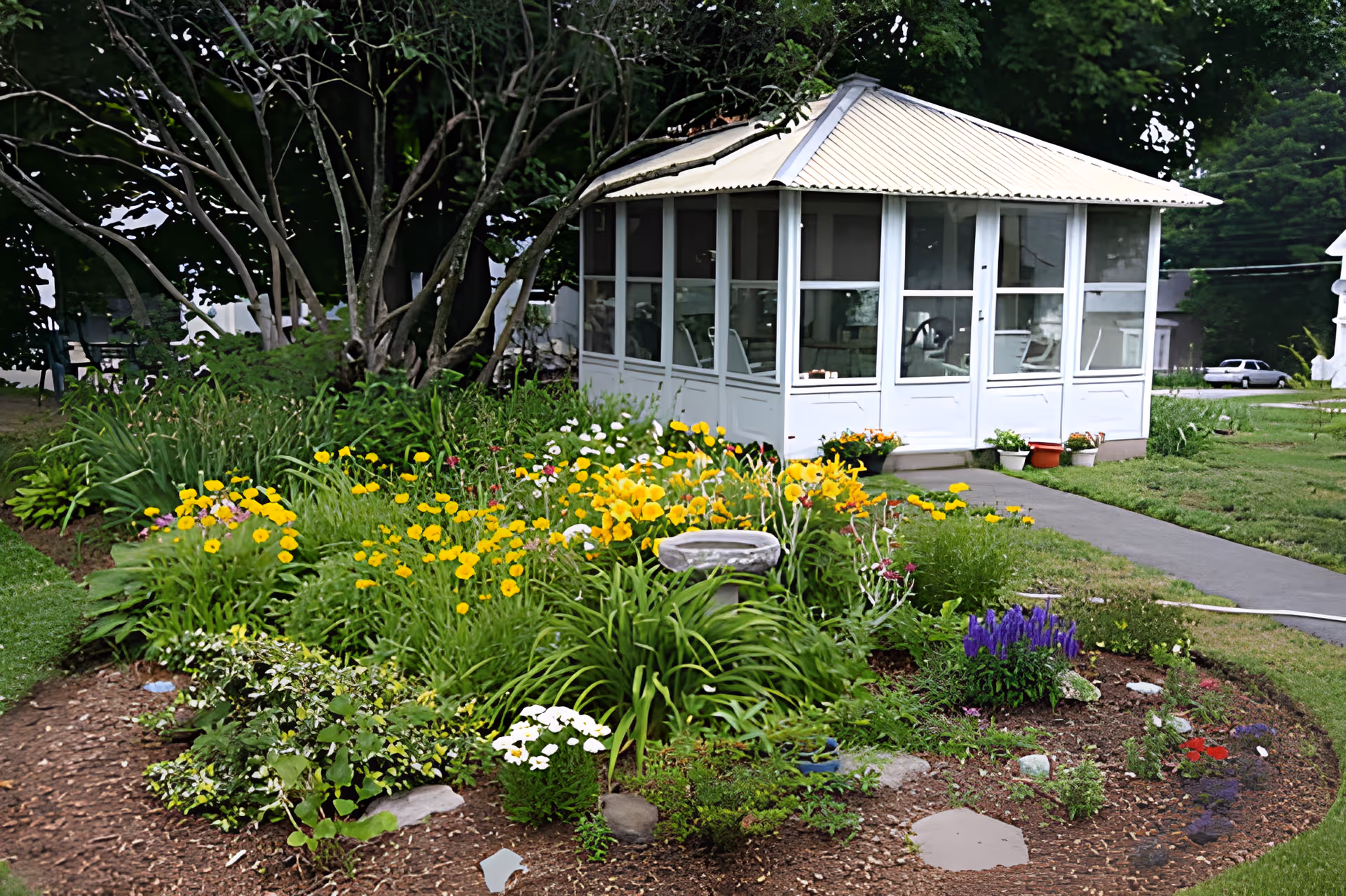 A vibrant garden with various colorful flowers including yellow, white, and purple blooms surrounding a small birdbath. Behind the garden is a white gazebo with large windows and a metal roof, situated on a grassy lawn with a paved walkway leading to it. Trees and other greenery are visible in the background.