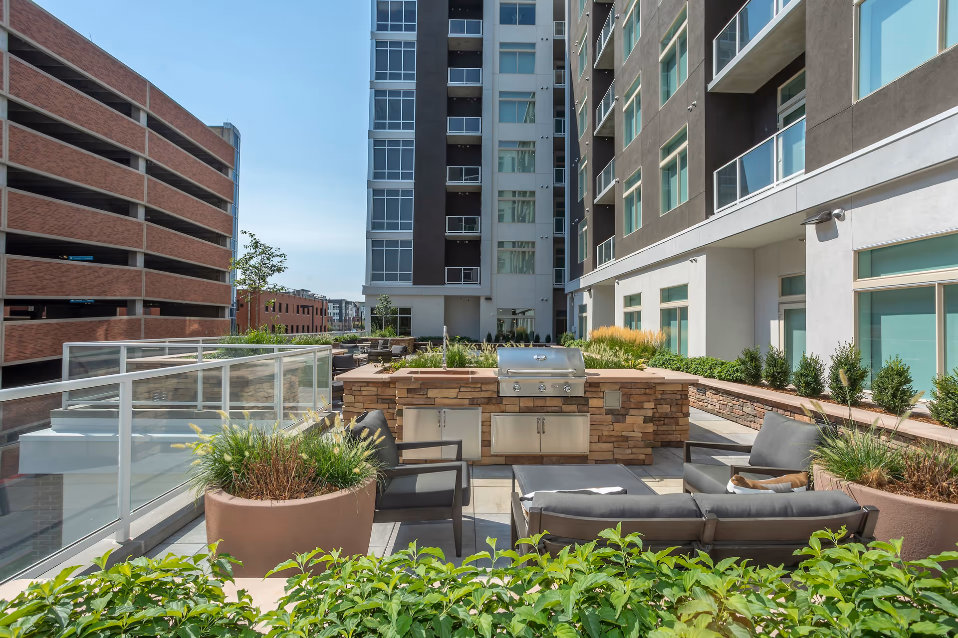 Outdoor patio area at Overture 9th + Co 55+ Apartment Homes featuring a built-in stone grill, modern outdoor seating with cushions, large planters with greenery, and a view of the apartment building and adjacent parking structure under a clear blue sky.