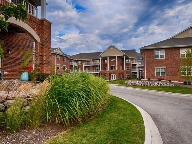 Exterior view of a red-brick senior living apartment complex with a curved driveway, landscaped grasses, and balconies under a cloudy sky.