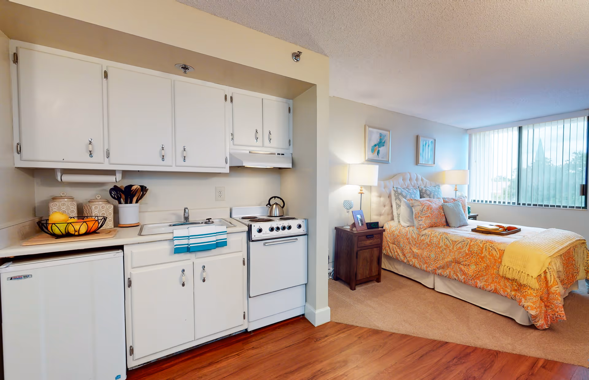 A small studio apartment featuring a compact kitchen area with white cabinets, a mini fridge, a stove, and a sink. Adjacent to the kitchen is a cozy bedroom area with a bed dressed in orange and white patterned bedding, two bedside tables with lamps, and a large window with vertical blinds letting in natural light.
