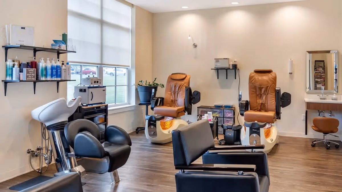 Interior view of a salon area in a senior living facility with two brown pedicure chairs, black salon chairs, a white hair washing station, shelves with hair care products, a window with blinds, and a small sink with a mirror and a brown stool.