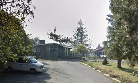 Exterior view of a single-story building with a covered porch, surrounded by trees and greenery. A white van is parked on the left side of the paved driveway leading up to the building.