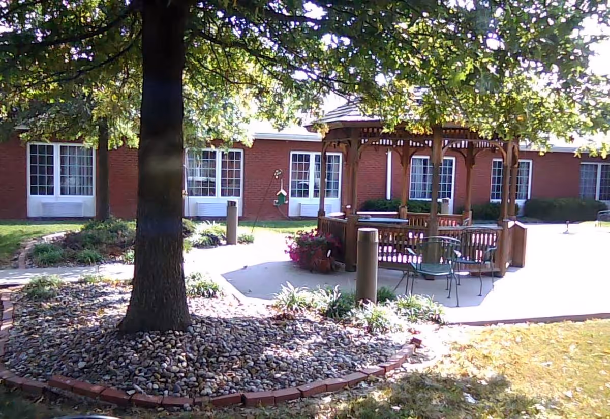 Outdoor courtyard area with a large tree surrounded by rocks and a brick border. There is a wooden gazebo with chairs underneath it, and a brick building with multiple windows in the background. The scene is shaded by tree leaves.