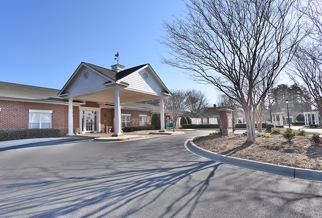 Exterior view of a single-story brick building with a covered entrance supported by white columns. There is a circular driveway in front, leafless trees, and a clear blue sky.