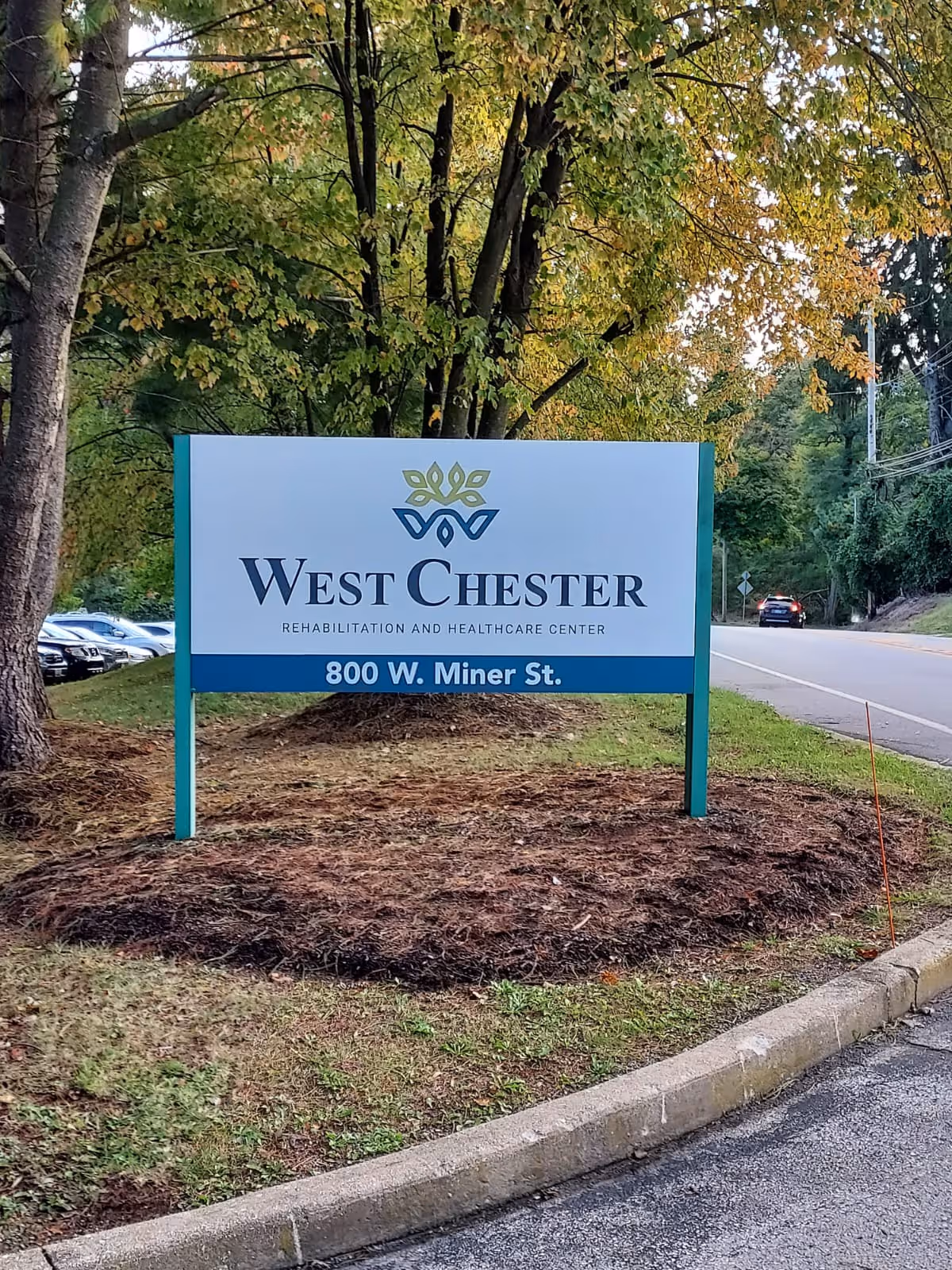 A freestanding roadside sign reading "West Chester Rehabilitation and Healthcare Center" with the address "800 W. Miner St." set by trees and a nearby road.