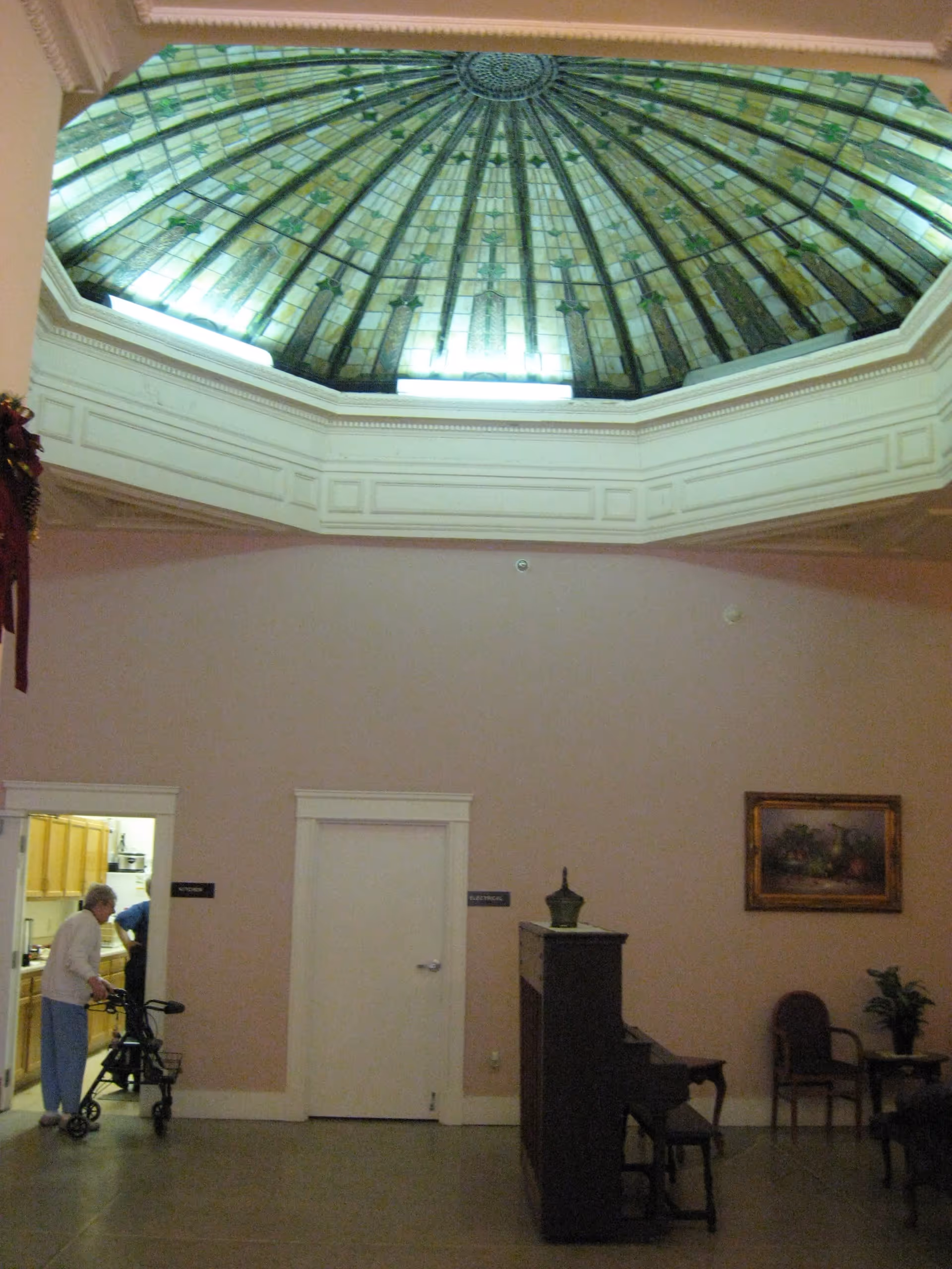 Interior view of a room with a large stained glass dome ceiling. An elderly person with a walker is near a doorway labeled 'Kitchen'. There is a piano against the wall, a framed painting, and a couple of chairs with a small table and plant.