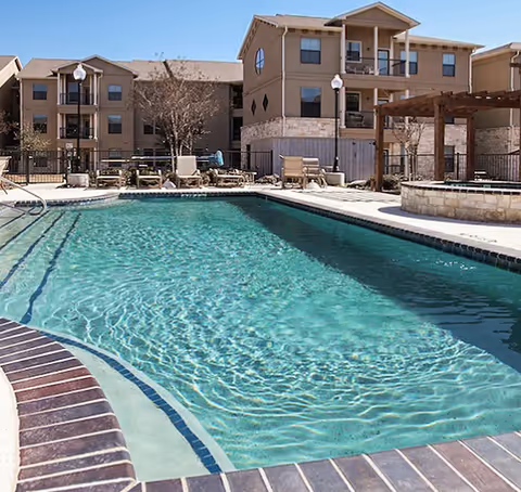 Outdoor swimming pool with lounge chairs and a three-story apartment building in the background.