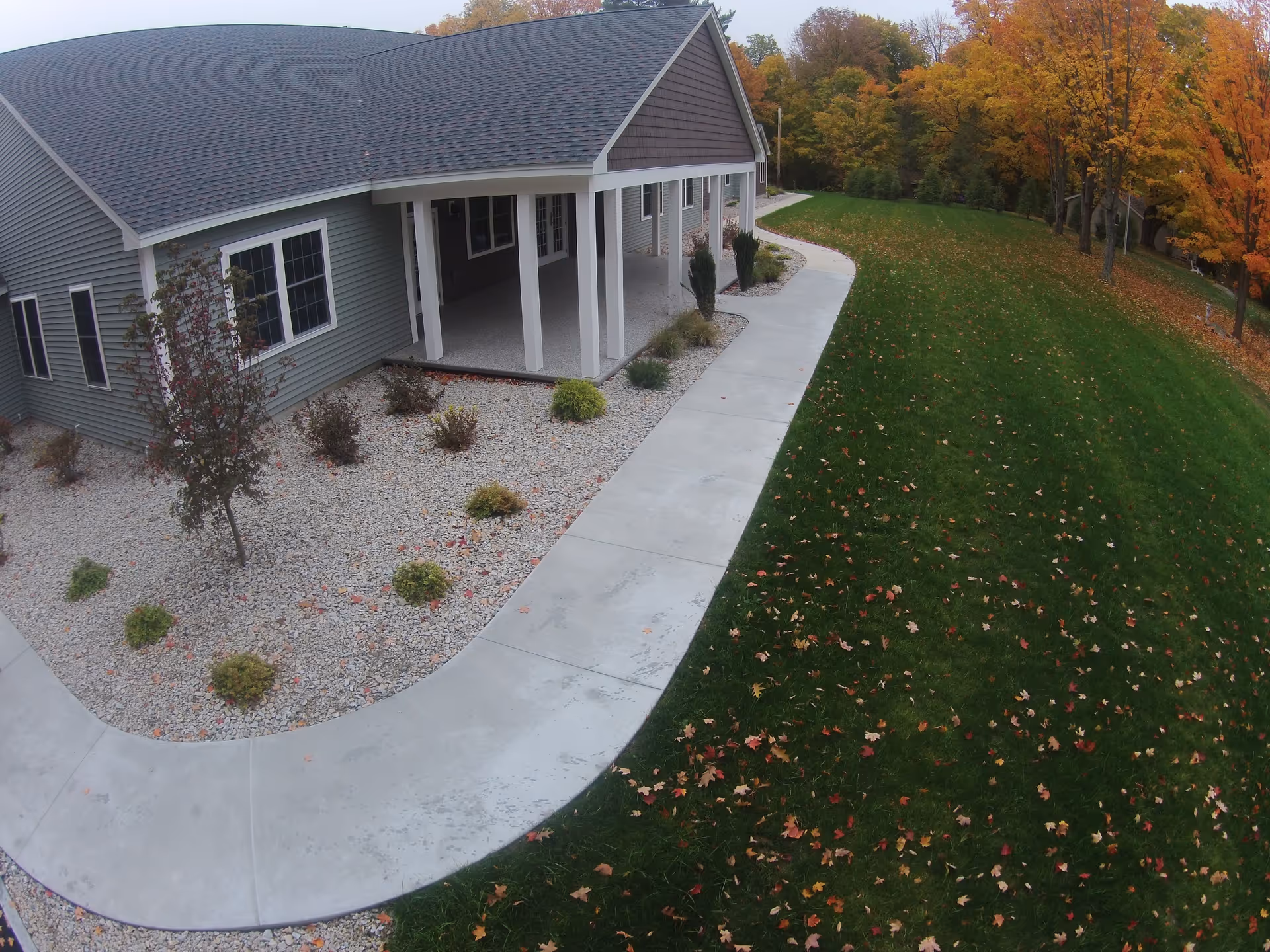 Exterior view of Beaver Lake Lodge Assisted Living showing a building with a covered porch, a concrete walkway, landscaped area with small bushes and trees, and a grassy lawn with scattered autumn leaves and trees with fall foliage in the background.