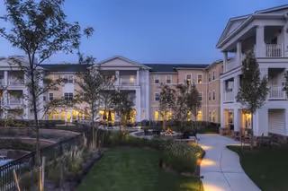Exterior view of a senior living facility building at dusk with multiple windows lit, surrounded by landscaped gardens, trees, and a paved walkway.