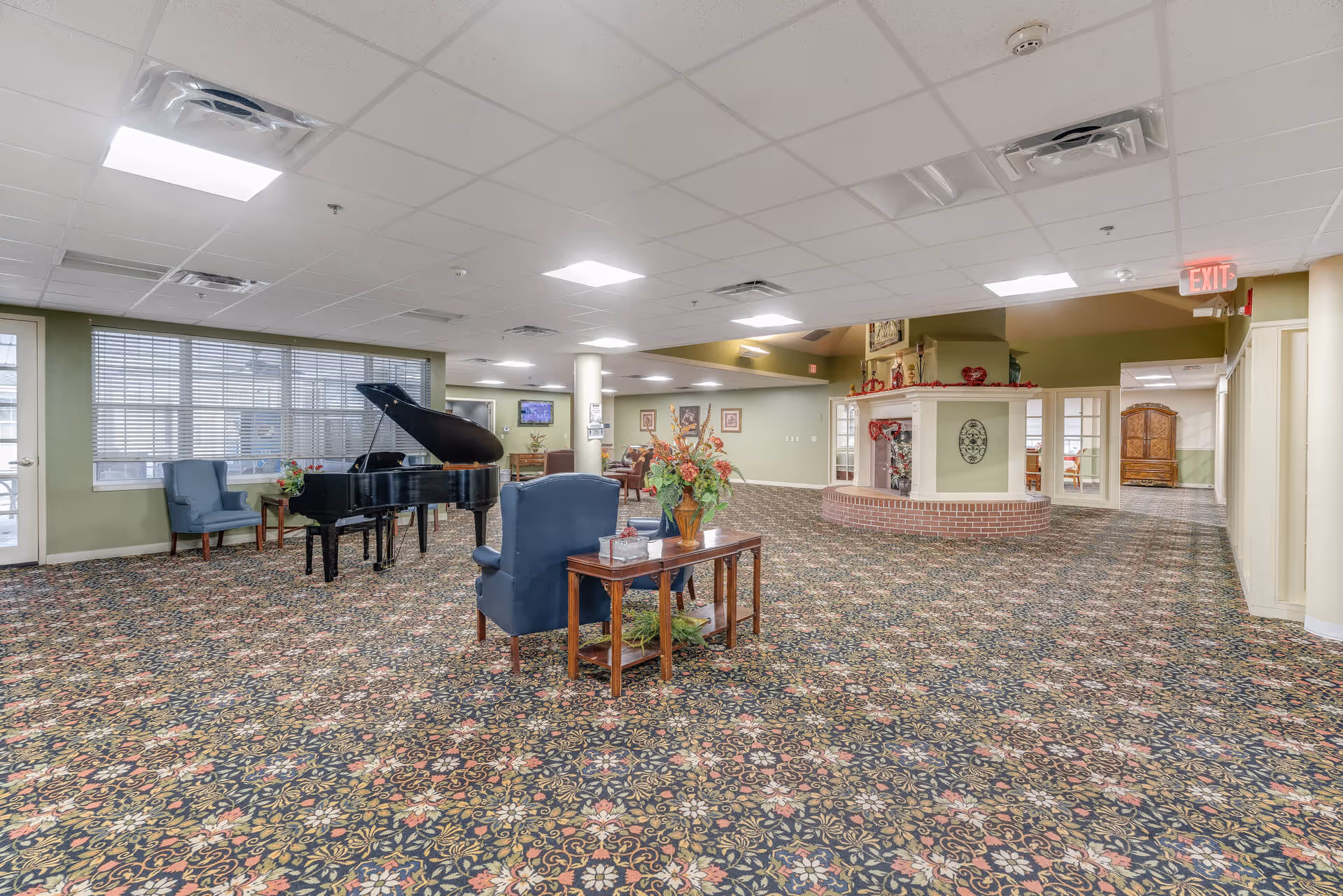 Spacious senior living common area with floral patterned carpet, a black grand piano, blue armchairs, a wooden side table with a flower arrangement, and a fireplace with decorative items. The room has a drop ceiling with recessed lighting and large windows with blinds.