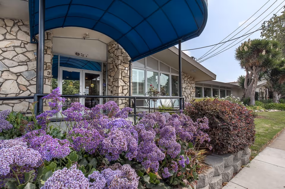 Stone-faced entrance of Windsor Gardens Rehabilitation Center with blue canopy and purple flowering shrubs.