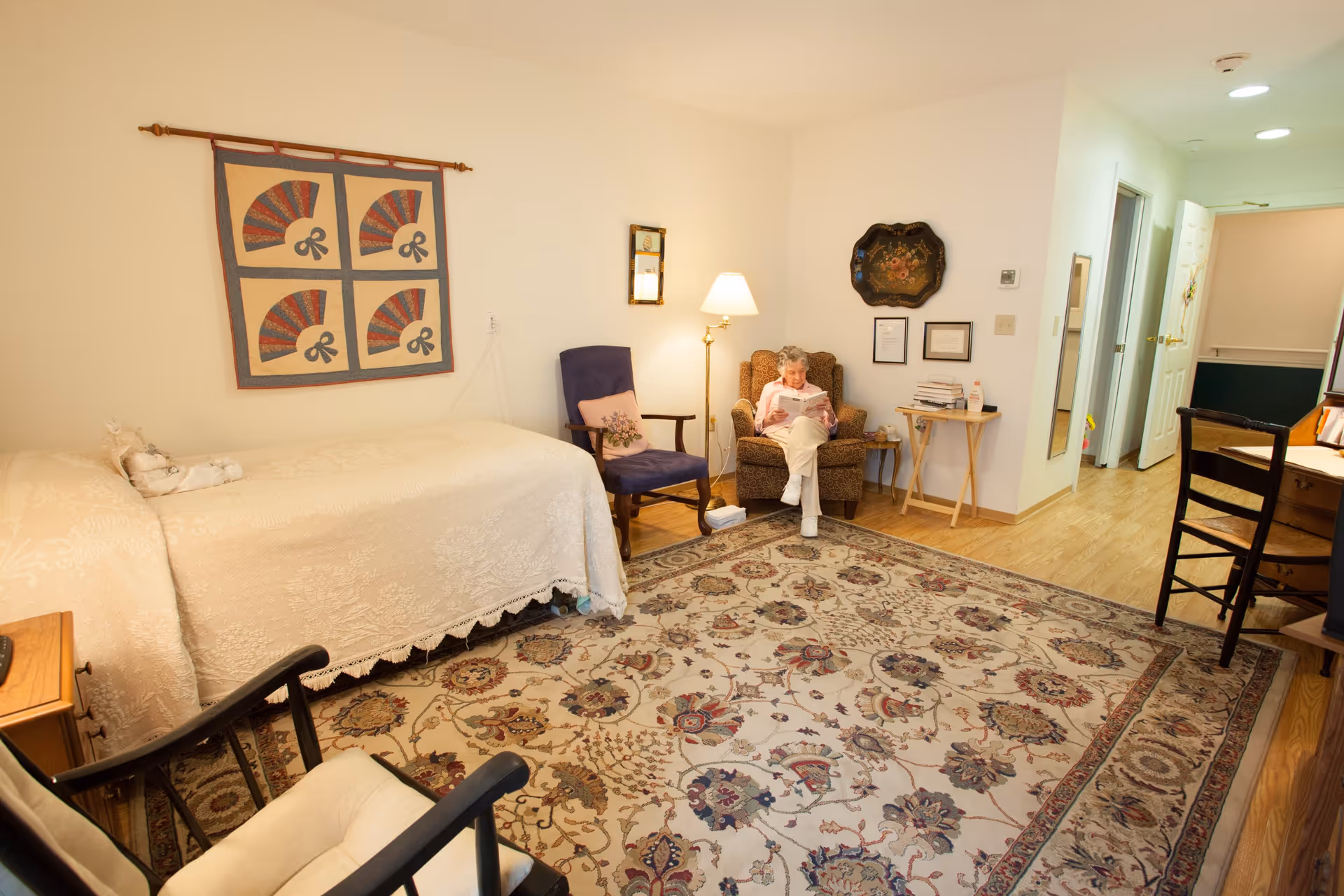 A cozy room with a single bed covered in a cream-colored bedspread, a large patterned area rug, and several chairs including a cushioned armchair where an elderly person is sitting and reading. The walls are decorated with a quilt and framed items, and there is a floor lamp providing warm light. The room has wooden flooring and an open doorway leading to a hallway.