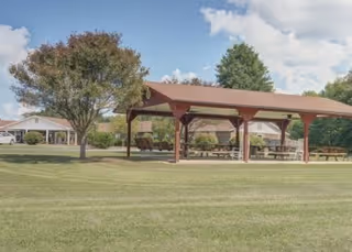 A large grassy outdoor area with a wooden pavilion featuring picnic tables underneath. Trees and a building are visible in the background under a partly cloudy sky.