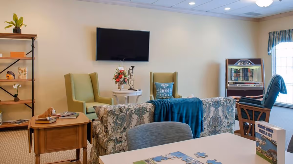 A cozy living room area with a floral patterned sofa draped with a blue blanket, two green armchairs, a wooden side table with books and a small dog figurine, a white round table with a flower vase and decorative items, a flat-screen TV mounted on the wall, a jukebox near a window with blue curtains, and a bookshelf with plants and decor.