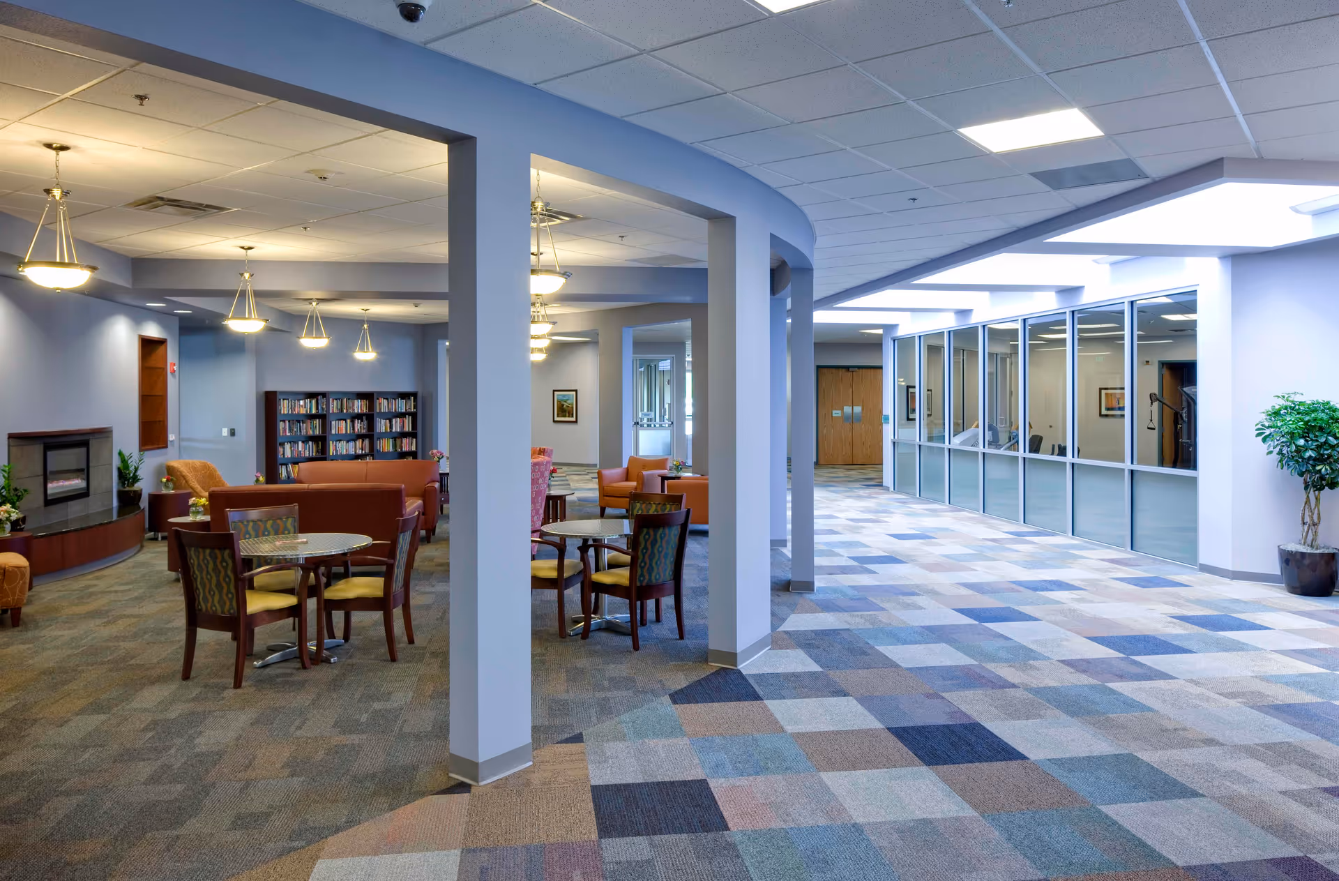 A spacious and well-lit common area in a senior living facility with multiple seating arrangements including round tables with chairs and sofas. There is a fireplace on the left side, bookshelves filled with books, and several ceiling lights. The floor has a patterned carpet, and there is a large windowed wall on the right side with a potted plant nearby.