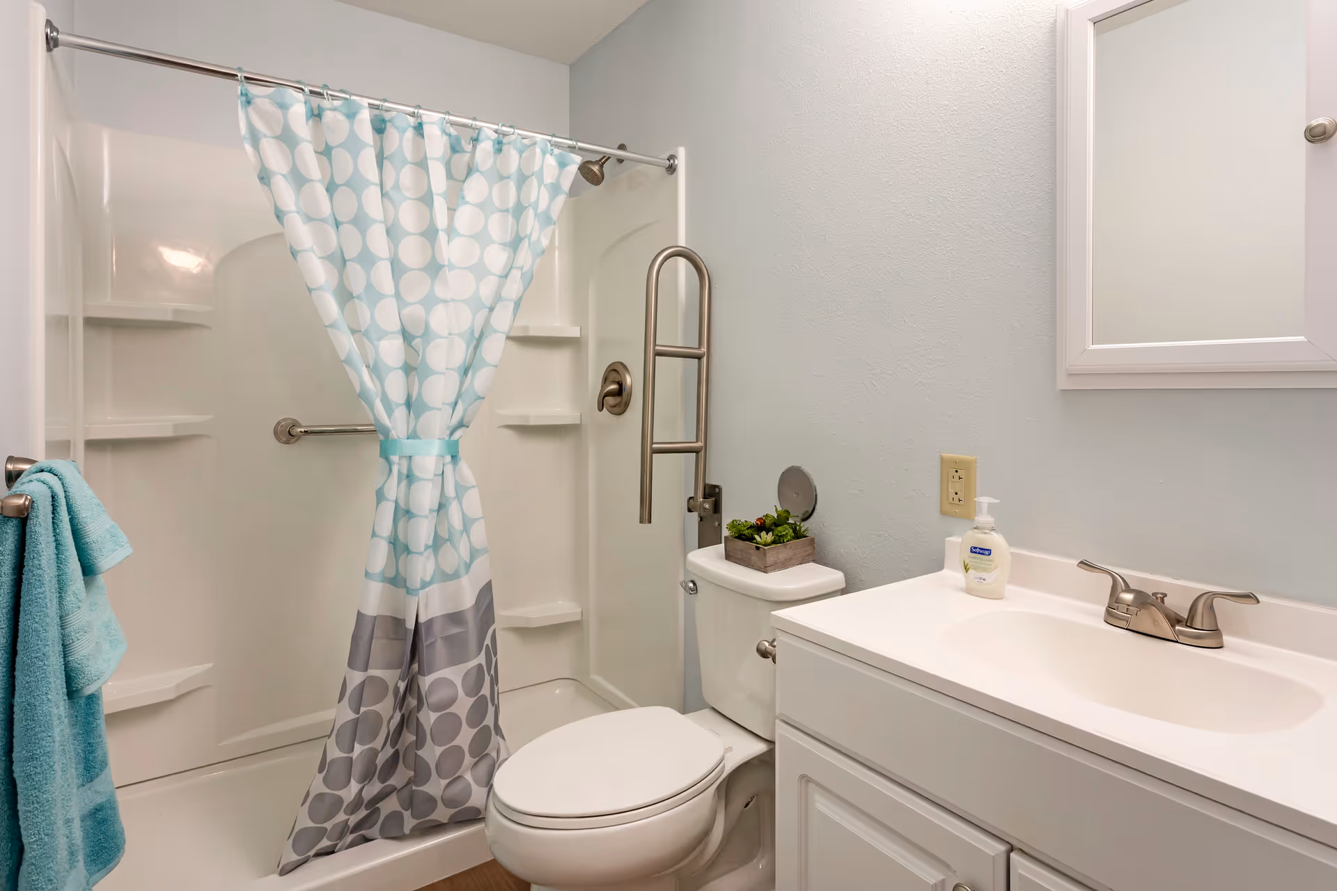A clean and simple bathroom featuring a white toilet with a small plant on top, a white vanity with a sink and faucet, a wall-mounted mirror cabinet, and a shower with a blue and gray polka dot curtain. There is a grab bar inside the shower and a towel rack with a folded turquoise towel on the left wall.