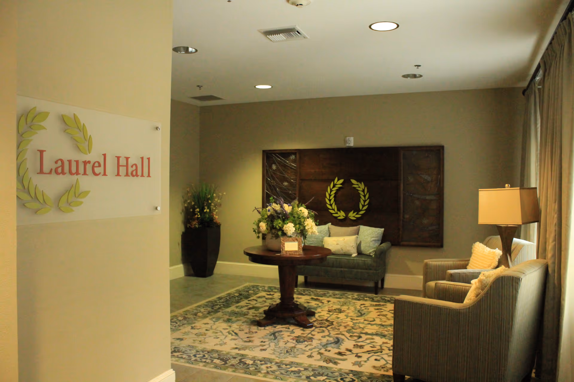 A cozy sitting area in Laurel Hall at The Wesleyan Assisted Living and Memory Care, featuring a round wooden table with a floral arrangement, a green cushioned loveseat with pillows, two striped armchairs with pillows, a floor lamp, and a decorative wall piece with a laurel wreath design. The area is softly lit with natural light coming through a window with curtains.