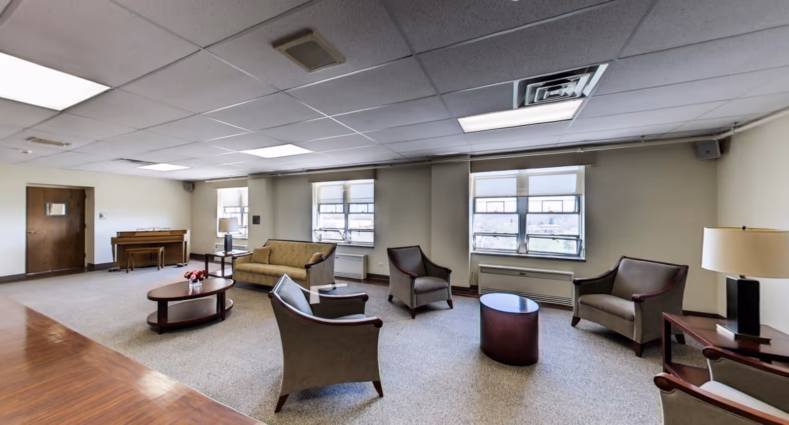 A spacious senior living common area with beige walls and carpeted floor. The room features a beige sofa, four armchairs arranged around two wooden coffee tables, a wooden piano against the far wall, and two windows with white blinds letting in natural light. There is a table lamp on a side table near one of the armchairs.