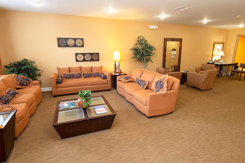 A spacious living room area with three orange sofas arranged around a dark wooden coffee table with glass inserts. The sofas have patterned cushions, and there are two potted plants and a table lamp providing warm lighting. The walls are painted beige, and there is wall art above one sofa and a large mirror on the far wall. In the background, there is a small table with chairs and additional seating.