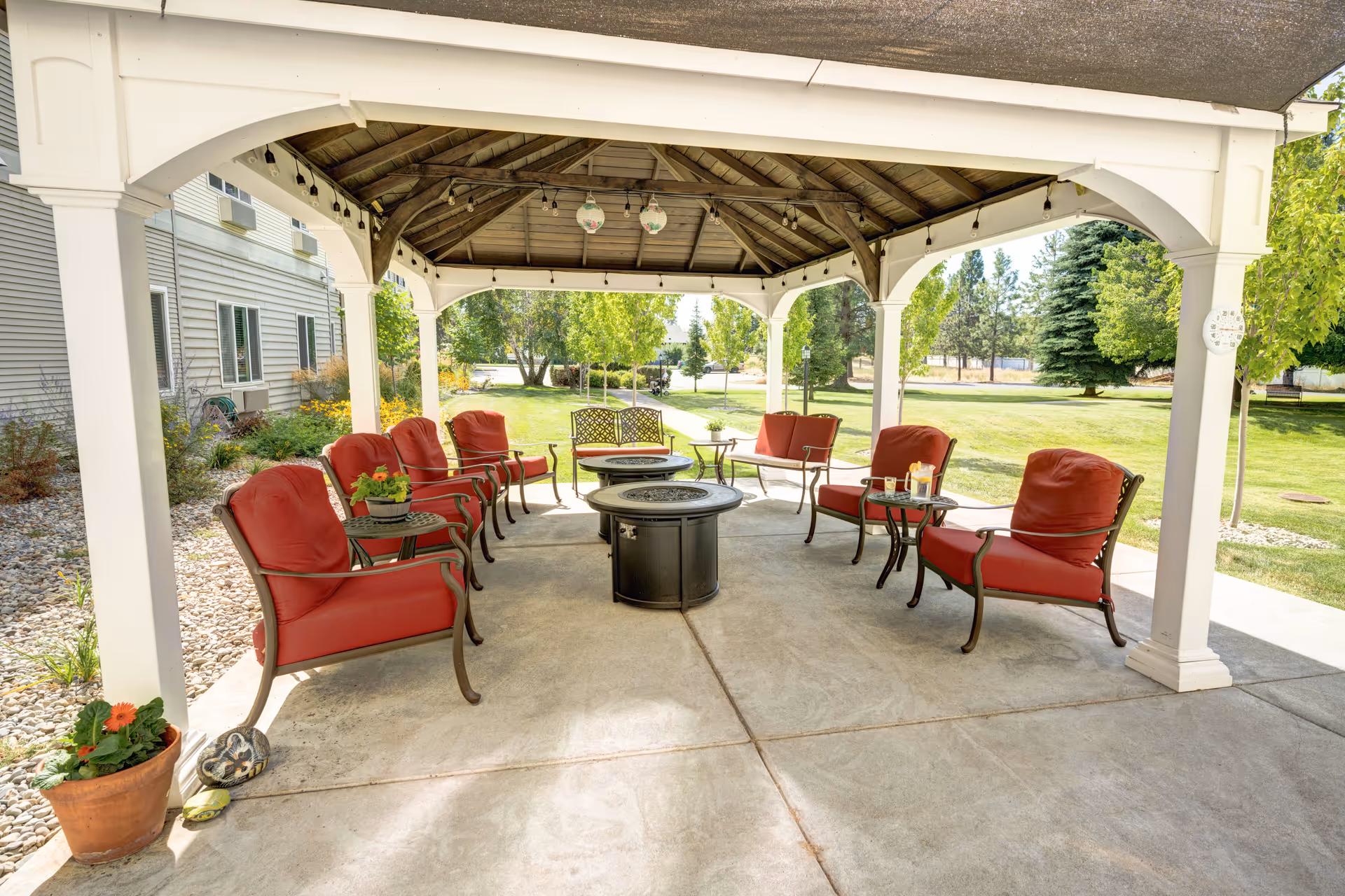 Outdoor covered seating area with red cushioned chairs arranged around two circular fire pits. The area is shaded by a wooden gazebo with string lights and hanging lanterns. Surrounding the gazebo is a well-maintained lawn with trees and shrubs, adjacent to a building with beige siding.
