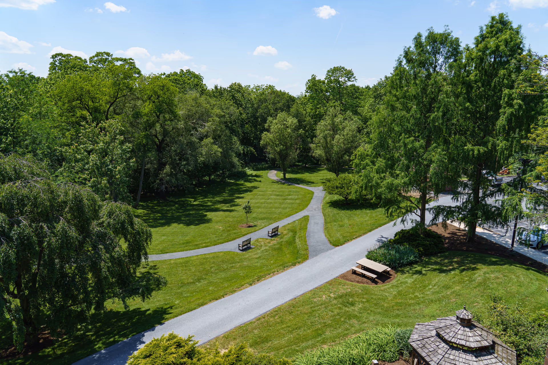 A scenic outdoor area at The Woods at Cedar Run featuring a well-maintained grassy lawn, several large green trees, a winding paved walking path, two benches along the path, a picnic table on the grass, and a small wooden gazebo in the lower right corner under a clear blue sky with a few clouds.
