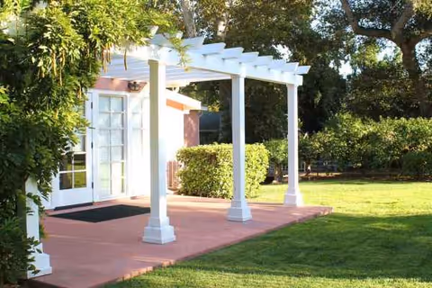White pergola over pink patio leads to glass-door entrance beside a green lawn.