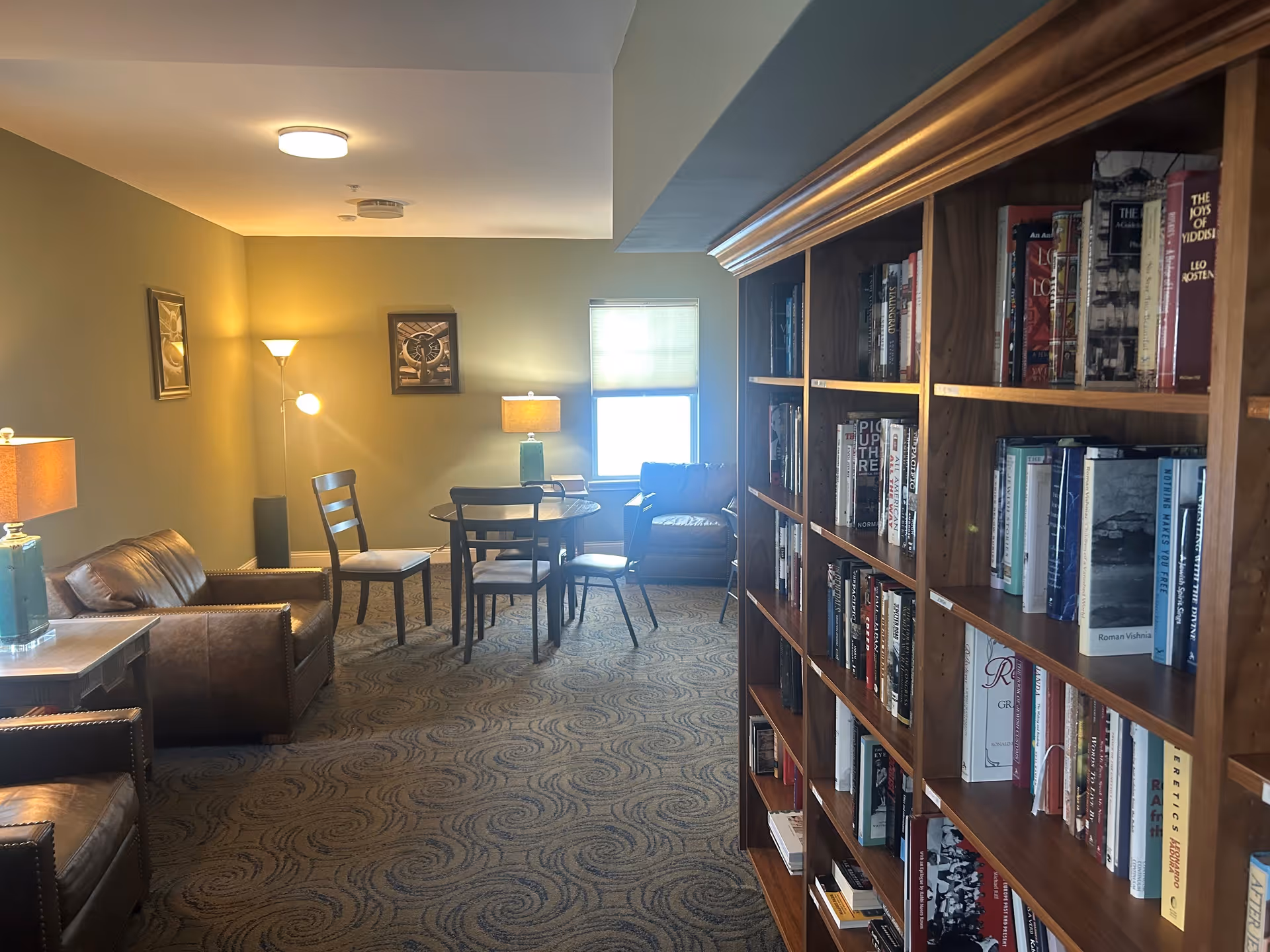 A cozy interior room with a large wooden bookshelf filled with books on the right side. The room features two brown leather armchairs, a round table with four chairs, two table lamps, and a floor lamp. The walls are painted light green, and there are framed pictures hanging on the wall. A window with a partially closed blind lets in natural light.