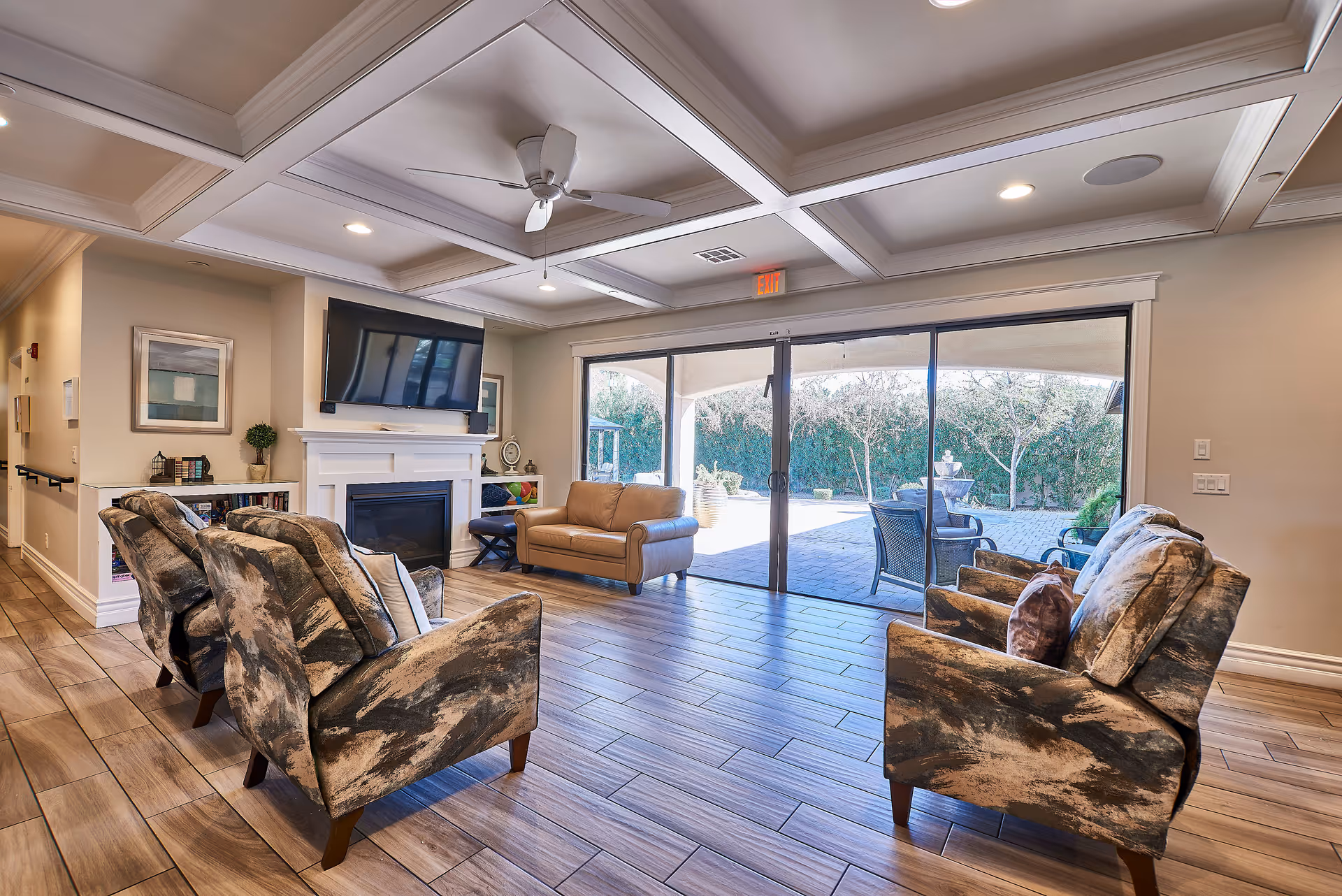 A bright and spacious living room with a coffered ceiling and ceiling fan. The room features a fireplace with a mounted flat-screen TV above it, a tan leather loveseat, and four patterned armchairs arranged around the fireplace. Large sliding glass doors open to an outdoor patio area with seating and greenery visible outside.