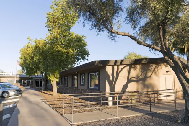 Exterior view of Desert Terrace Healthcare Center showing a single-story building with several windows, a wheelchair accessible ramp, trees, and parked cars along a sidewalk under a clear sky.