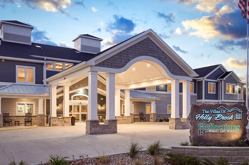Front entrance of the Villas of Holly Brook assisted living building with a covered porte-cochère and an illuminated sign at dusk.