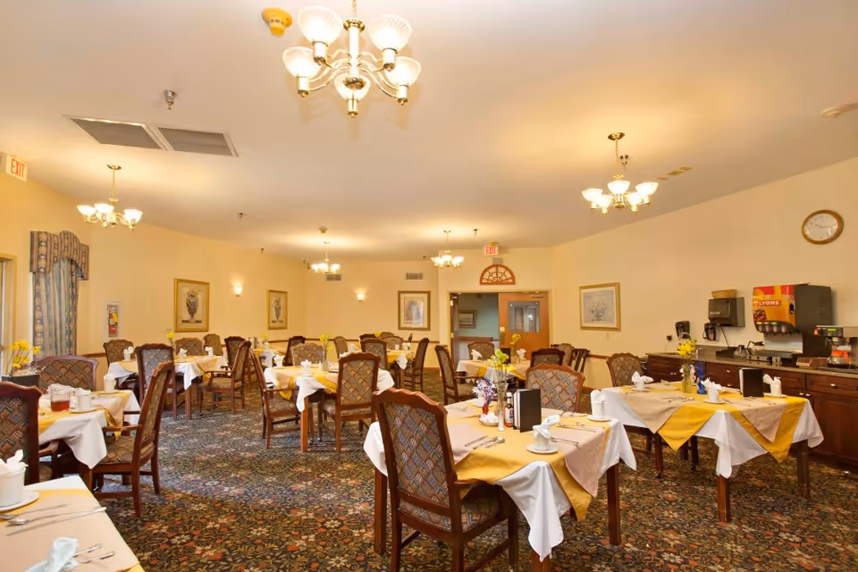 A well-lit dining room with multiple tables covered in white and yellow tablecloths, each set with cups, napkins, and utensils. The room features patterned carpet, upholstered chairs, chandeliers, framed artwork on the walls, and a beverage station with a soda dispenser and coffee maker along one wall.