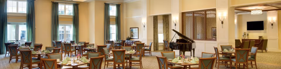 Elegant dining room with multiple set tables, tall windows with green curtains, and a grand piano near a seating area.